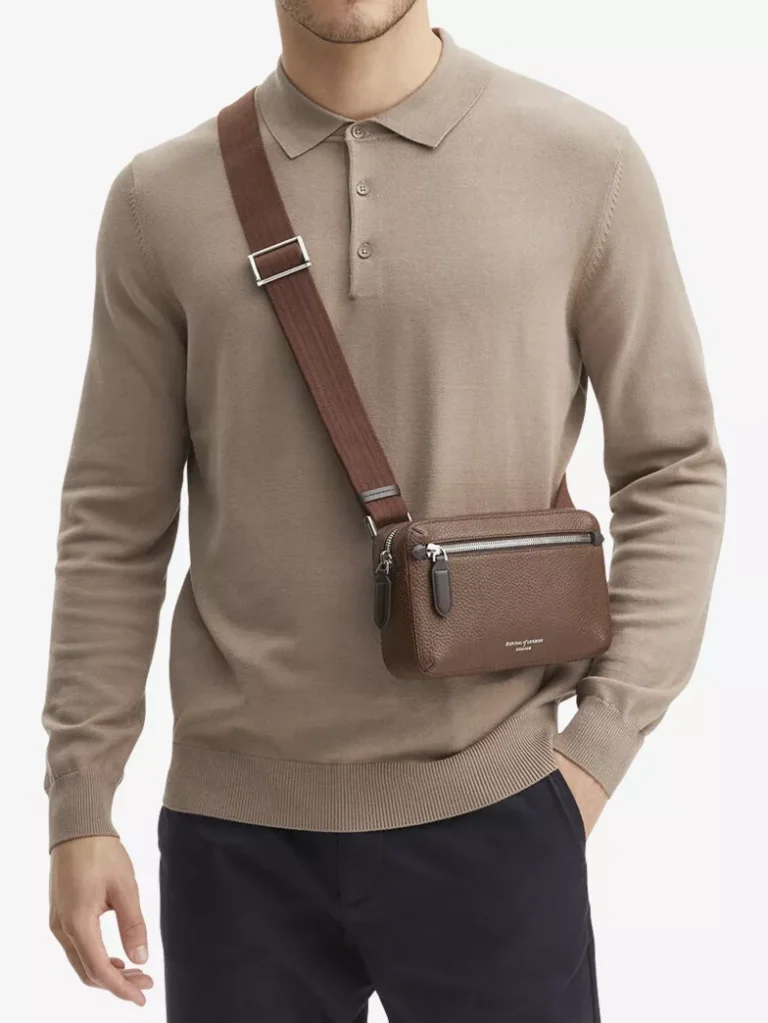 Torso of a male model wearing a brown Aspinal of London grained-leather messenger bag across the chest, styled with a beige long-sleeve knit polo and navy trousers against a white background.