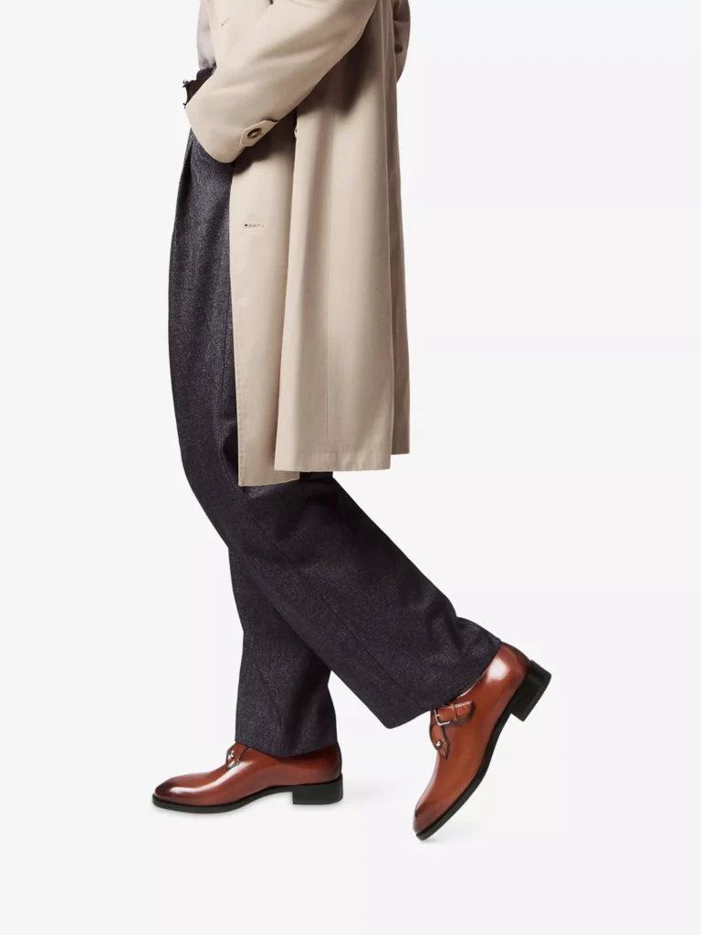 Lower half of a male model wearing Christian Louboutin brown calf-leather Chambelimonk shoes, styled with dark charcoal trousers and a beige trench coat against a white background.