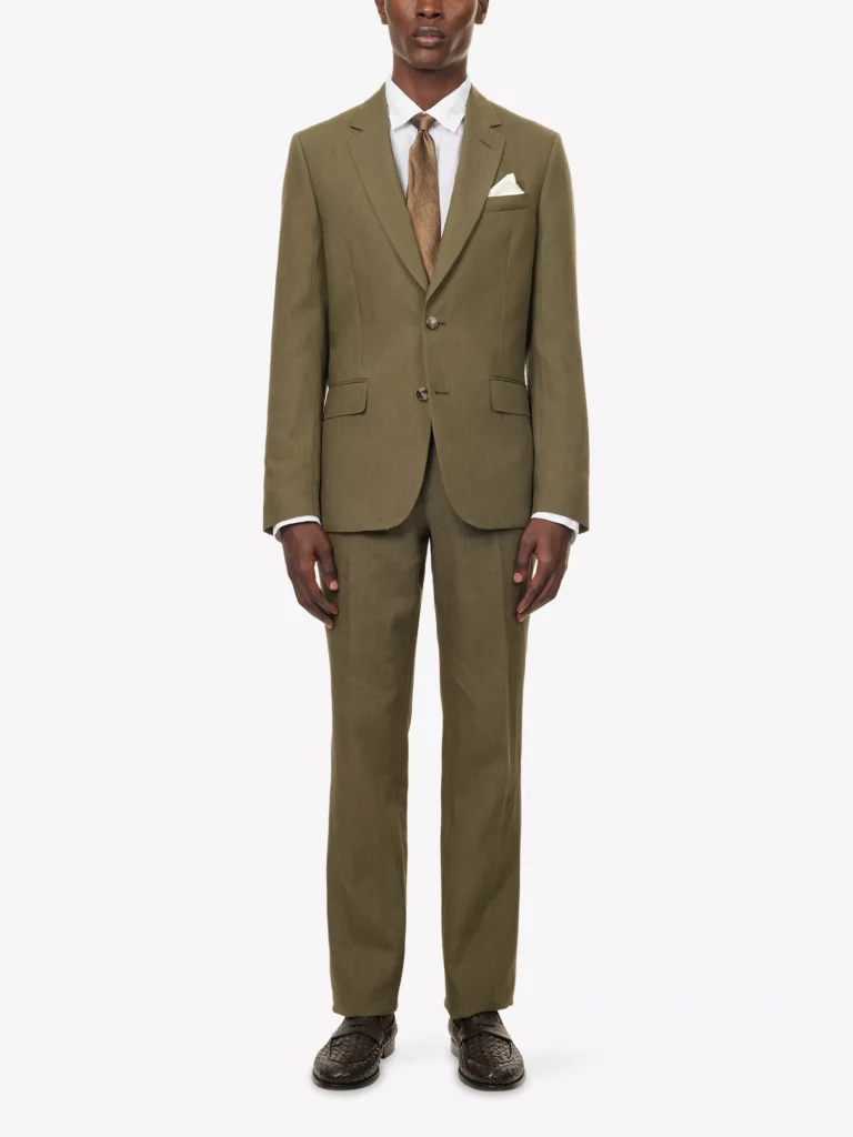 Torso of a male model wearing an olive green soft tailored suit, styled with a white shirt, textured tan tie, white pocket square, and dark woven loafers against a white background.