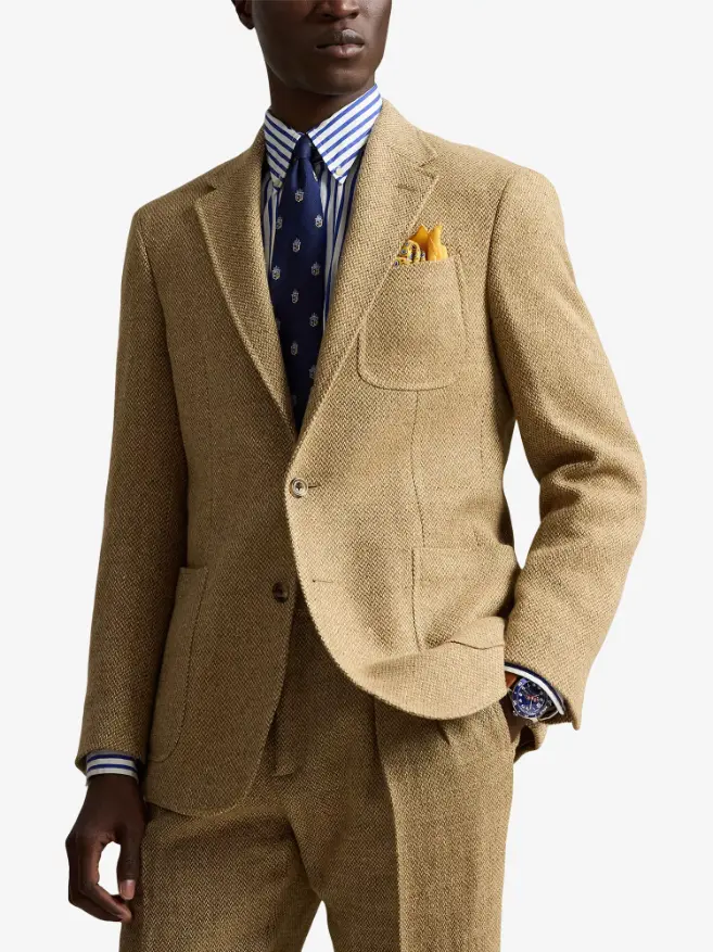 Torso of a male model wearing a tan Polo Ralph Lauren summer tweed blazer, styled with a blue and white striped shirt, navy patterned tie, and a yellow pocket square against a white background.